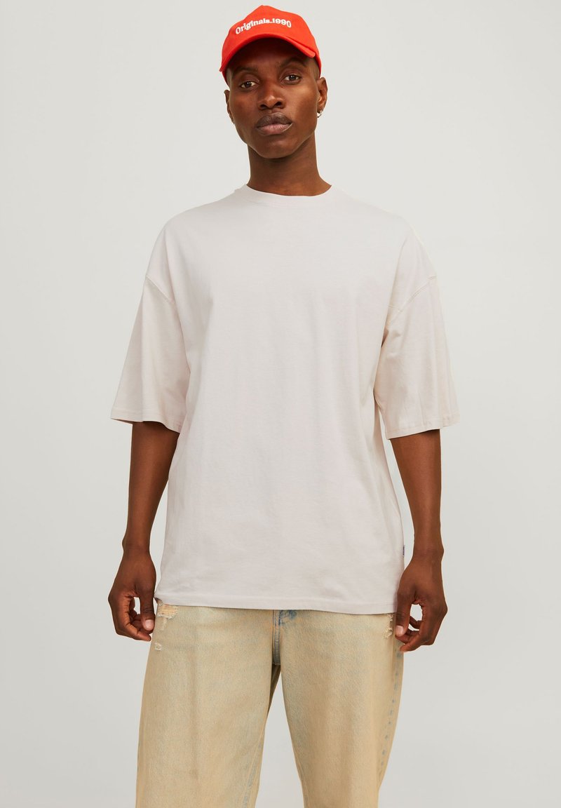 Lys beige oversized T-skjorte med korte ermer, laget av bomull, kombinert med lyseblå slitte jeans og en lys rød baseball caps.