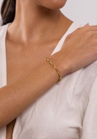 Goldene Kettenarmband mit abwechselnd ovalen und runden Gliedern, das mit einer polierten Oberfläche versehen ist. Es wird an einem nackten Handgelenk über einem hellen Stoff getragen.