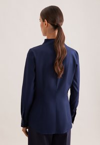 Navy blue blouse featuring a stand collar, long sleeves with button accents, and a fitted silhouette with a smooth texture.