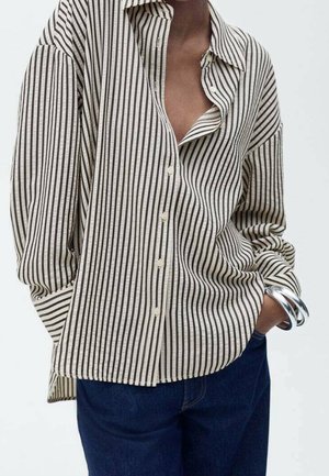 Persona que lleva una camisa beige holgada de manga larga con rayas negras verticales, jeans azul oscuro y pulseras plateadas en la muñeca.