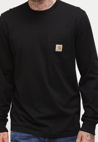 Černá dlouhá rukávová košile vyrobená z bavlny, s kapsou na hrudi a štítkem Carhartt. Hladký povrch, kulatý výstřih.
