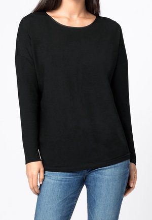 Kobieta ubrana w jednolity czarny długi rękaw i jasnoniebieskie jeansy, stojąca na białym tle.