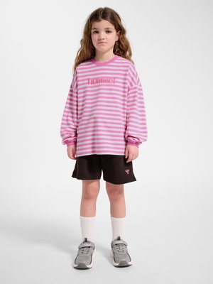 Mädchen steht und trägt ein rosa-weiß gestreiftes Langarmshirt, schwarze Shorts, weiße Socken und graue Sneakers vor einem schlichten Hintergrund.