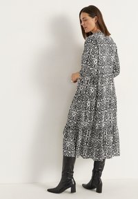 Langes, schwarz-weiß gemustertes Kleid mit langen Ärmeln und gestuftem Saum, kombiniert mit schwarzen knielangen Stiefeln. Glattes, fließendes Material.