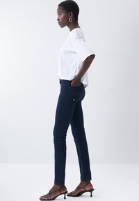 Jean skinny bleu foncé avec une taille mi-haute, fabriqué en denim extensible. Accompagné d'un haut blanc ample et de talons noirs à lanières.