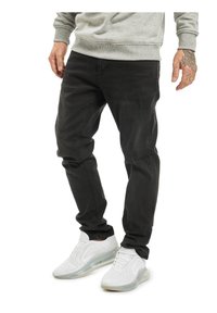 Muž nosící černé potrhané džíny, bílé tenisky s průhlednými podrážkami a šedou mikinu, stojící s viditelnými tetovanými rukama.