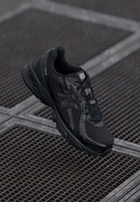 Chaussure de sport noire avec des panneaux en mesh, semelle en caoutchouc et lacets. Présente un logo réfléchissant et un dessus texturé pour l'adhérence et le soutien.
