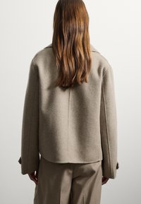 Abrigo de lana beige con un corte relajado, que cuenta con un cuello, costuras visibles y puños abotonados, combinado con pantalones beige.