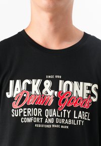 En nærbilde av en persons nakke og bryst som har på seg en svart T-skjorte med teksten "JACK & JONES Denim Goods" i fete, fargerike bokstaver.