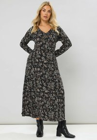 Lungo vestito nero con motivo floreale nelle tonalità crema e marrone, caratterizzato da un scollo a V e maniche lunghe. Indossato con stivaletti neri con tacco.