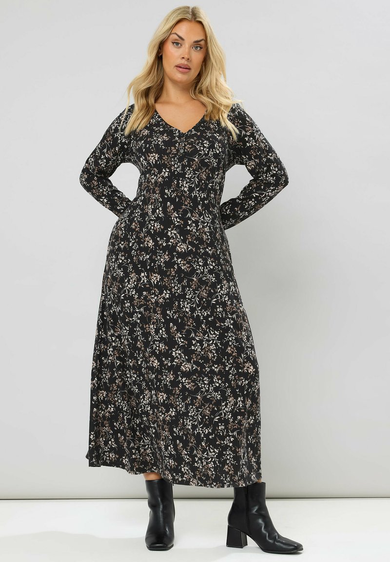 Lungo vestito nero con motivo floreale nelle tonalità crema e marrone, caratterizzato da un scollo a V e maniche lunghe. Indossato con stivaletti neri con tacco.