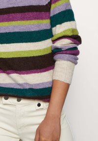 Bunter Pullover mit Streifen in Grün, Lila, Braun und Creme; gerippte Bündchen und Saum; weiche Textur. Kombiniert mit hellfarbigen Hosen.