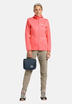 Frau mit grauen Haaren, die eine korallenfarbene Jacke, beige Hosen und Wanderschuhe trägt und eine kleine marineblaue Reisetasche vor einfarbigem Hintergrund hält.