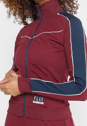 Femme portant une veste zippée bordeaux avec des rayures bleu marine et blanches sur les manches et une étiquette "PLAY EST. 2006" près de la taille.