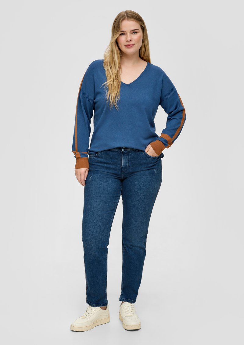 Suéter de punto azul con escote en V, mangas largas con rayas marrones, combinado con jeans oscuros y zapatillas blancas.