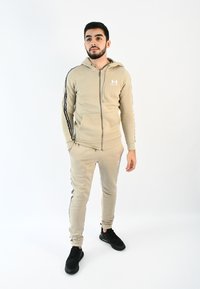 Chándal deportivo beige con sudadera con cremallera, rayas laterales negras y logo. Combinado con zapatillas negras. Tejido suave; diseño ajustado.