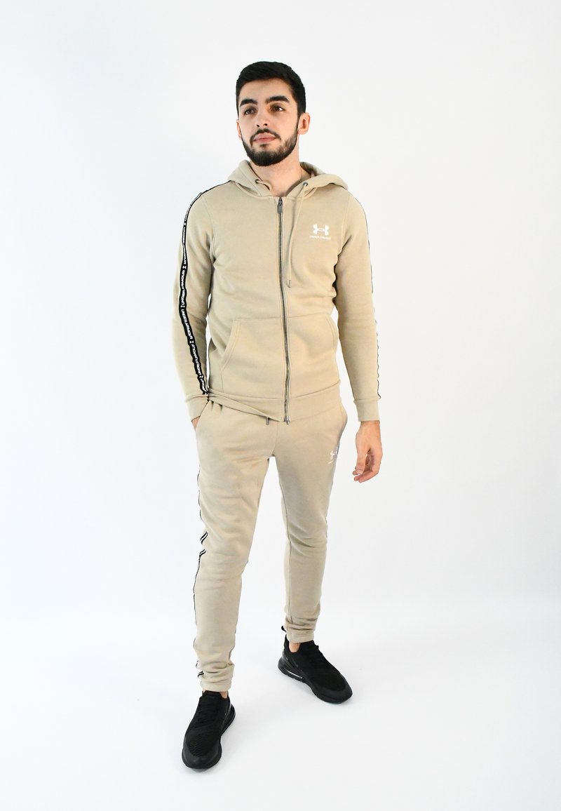 Chándal deportivo beige con sudadera con cremallera, rayas laterales negras y logo. Combinado con zapatillas negras. Tejido suave; diseño ajustado.