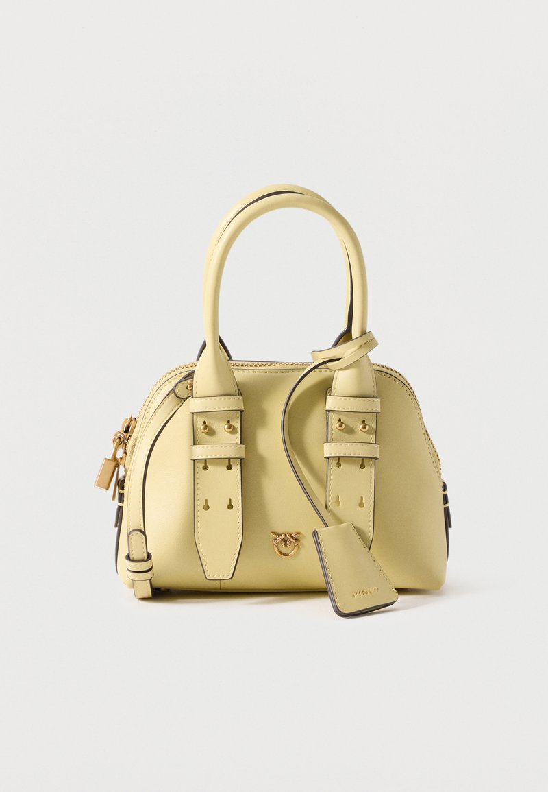 Μικρή δερμάτινη τσάντα σε ανοιχτό κίτρινο χρώμα με δύο λαβές, διακοσμητικά αγκράφες, χρυσό λογότυπο και κρεμαστή δερμάτινη ετικέτα σε ανοιχτό φόντο.