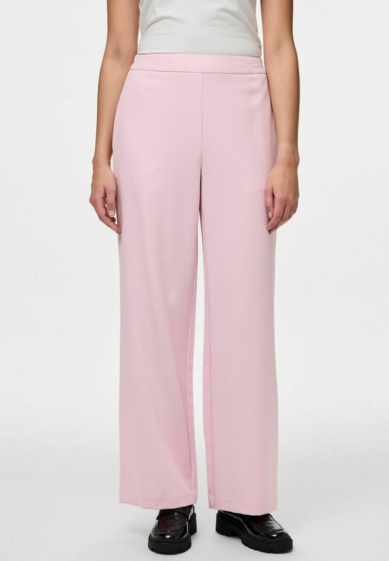 Pantalons larges rose clair en tissu doux avec une texture lisse, dotés d'une taille haute et de poches latérales. Des chaussures noires complètent le look.