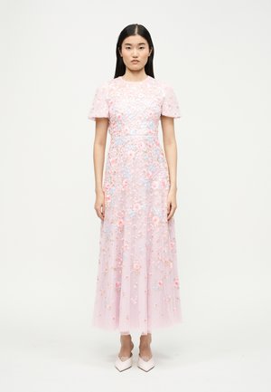 Femme debout portant une robe longue rose pâle à manches courtes avec une broderie florale multicolore, assortie à des chaussures pointues rose clair.