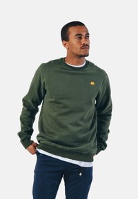 Grøn sweatshirt med et struktureret stof, rund halsudskæring, ribbede manchetter og et lille gult elefantlogo på brystet. Bæret over en hvid t-shirt.