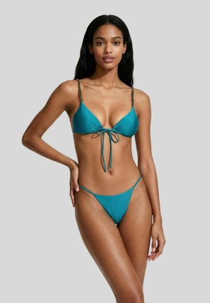 Donna in piedi che indossa un bikini verde acqua a due pezzi con spalline a catena sulla parte superiore, posando davanti a uno sfondo grigio chiaro uniforme.