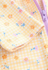 Dzeltena auduma ar šaha rakstu un ziedu motīvu, kas rotāts ar rozā, oranžām un zilām ziedu krāsām. Priekšpusē ir lavandas krāsas rāvējslēdzis. Mīksta tekstūra.