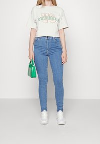 Kvinna klädd i en vit grafisk t-shirt, blå smala jeans, vita sneakers och håller en liten grön handväska vid sin sida.