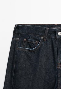 Dunkelblaue Denim-Jeans mit kontrastierenden orangefarbenen Nähten, mit einem Knopfverschluss, Vordertasche und subtiler Textur. Das Etikett trägt die Aufschrift "Massimo Dutti."