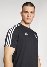 Sort atletisk T-shirt med hvidt tre-stribe design på skuldrene og et lille logo på brystet. Korte ærmer og rund halsudskæring.