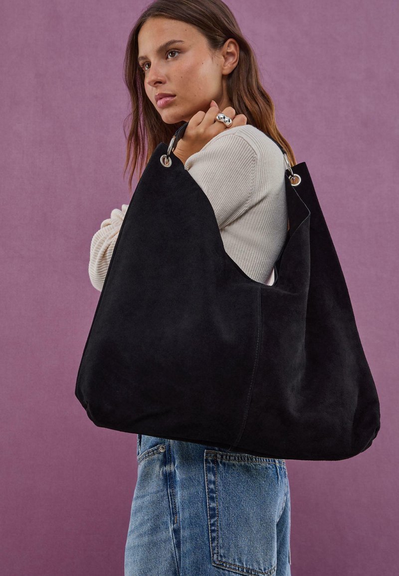 Sac hobo en suede noir avec une forme décontractée, des accents en métal argenté et un dessus ouvert. Surface texturée avec une finition douce.