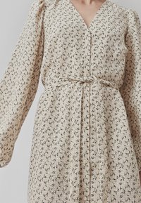 Beiges Kleid mit V-Ausschnitt, langen puffärmel, Knopfverschluss und Bindegürtel. Es hat ein florales Muster in Schwarz auf einem strukturierten Stoff.