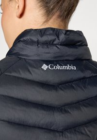 Crni puffer prsluk s chevron kvadratnim dizajnom, sjajni najlon, visok ovratnik i bijeli Columbia logo na leđima.