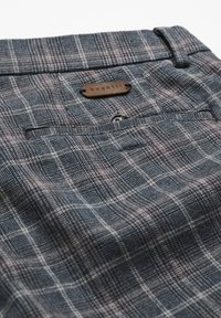 Tissu à carreaux gris et rouge avec une poche arrière comportant un bouton et une petite étiquette en cuir marron cousue au-dessus de la poche.