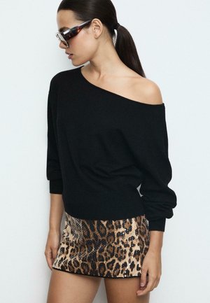 Schwarzer schulterfreier Pullover, kombiniert mit einem metallischen Minirock im Leopardenmuster, der Pailletten und eine taillierte Passform aufweist. Klare Sonnenbrillen runden den Look ab.