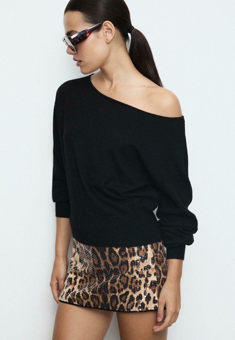 Schwarzer schulterfreier Pullover, kombiniert mit einem metallischen Minirock im Leopardenmuster, der Pailletten und eine taillierte Passform aufweist. Klare Sonnenbrillen runden den Look ab.