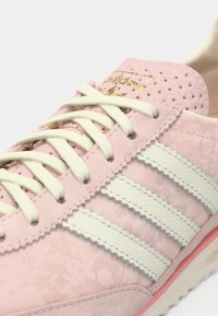 Rózsaszín sportcipő texturált felsőrésszel, fehér háromsávos mintával és világos fűzőkkel. Finom mintázatokkal és arany márkajelzéssel díszítve.