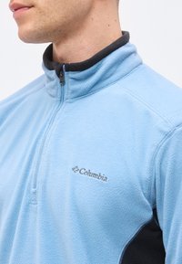 Άνδρας που φορά ελαφρύ γαλάζιο fleece μπουφάν Columbia με μαύρο γιακά και φερμουάρ μέχρι τη μέση, ορατό λογότυπο στο στήθος πάνω σε μονόχρωμο φόντο.