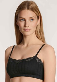 Schwarzer Bralette aus weichem Material, mit Spitzenbesatz entlang des Ausschnitts, vertikaler Rippstruktur und verstellbaren dünnen Trägern.