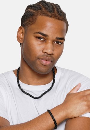 Ensemble de collier et bracelet en perles noires, présentant un design torsadé, porté sur un t-shirt blanc. Texture lisse avec une finition brillante.