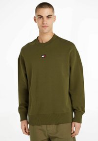 Sudadera verde oliva hecha de una mezcla de algodón. Presenta un cuello redondo, puños acanalados y un pequeño parche con el logo en el pecho.