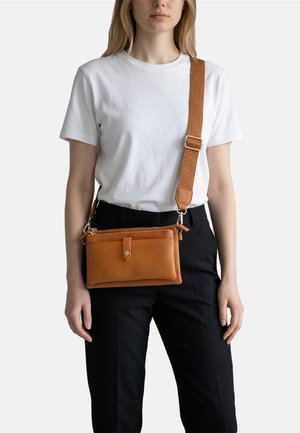 Donna che indossa una maglietta bianca e pantaloni neri, con una borsa a tracolla in pelle marrone con tracolla regolabile.