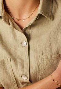 Gouden ketting met kleine geometrische bedeltjes, gedragen over een beige gestructureerd shirt met een zilveren knoop en zakdetail.