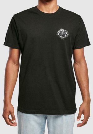 Homme portant un t-shirt noir à manches courtes avec un petit motif de rose blanche sur la poitrine gauche, associé à un jean en denim bleu clair.