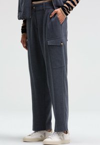 Dunkelgraue, maßgeschneiderte Hose mit lockerer Passform, ausgestattet mit einer seitlichen Cargo-Tasche und Messingknopf-Details. Getragen mit weißen Sneakers.