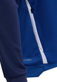 Haut de sport long à manches bleu en tissu texturé, avec une fermeture éclair blanche sur le côté et le logo "BE COOL" sur l'ourlet. Présente un poignet de manche contrastant.