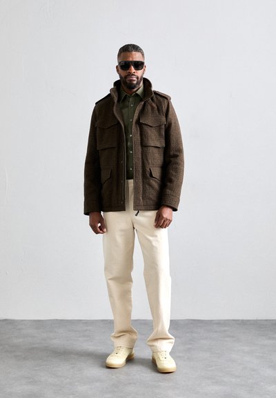 Veste marron texturée avec quatre poches avant, chemise verte en dessous, pantalon crème et baskets jaunes. Modèle portant des lunettes de soleil noires.