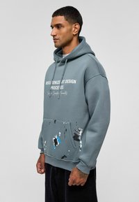 Grijze hoodie met een voorzak en verfspetters in blauw, zwart en wit, en witte tekst op de borst. Zacht materiaal.