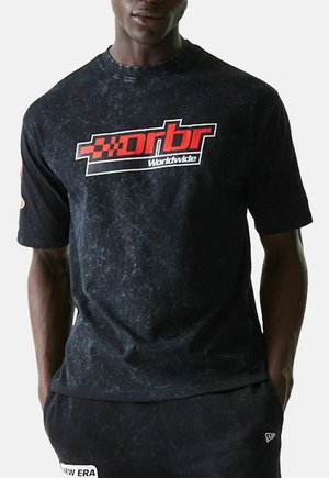 Mann trägt ein schwarzes, marmoriertes T-Shirt mit kurzen Ärmeln und rotem sowie weißem "drbr Worldwide"-Logo, kombiniert mit schwarzen Hosen mit weißem "New Era"-Aufnäher.