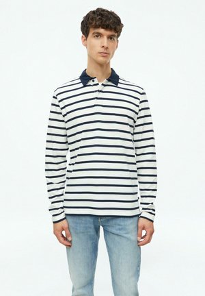 Junger Mann steht, trägt ein weißes Langarmshirt mit marineblauen horizontalen Streifen und einem dunklen Kragen, kombiniert mit hellblauen Jeans.